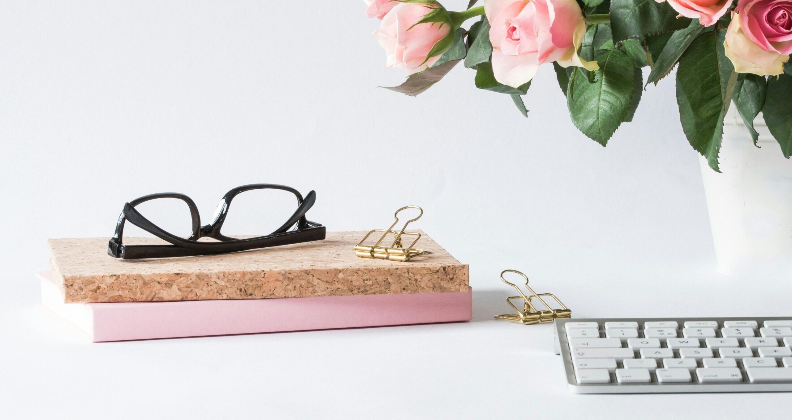Stylish workspace featuring pink roses, glasses, keyboard, and notebooks. Perfect for spring office decor.